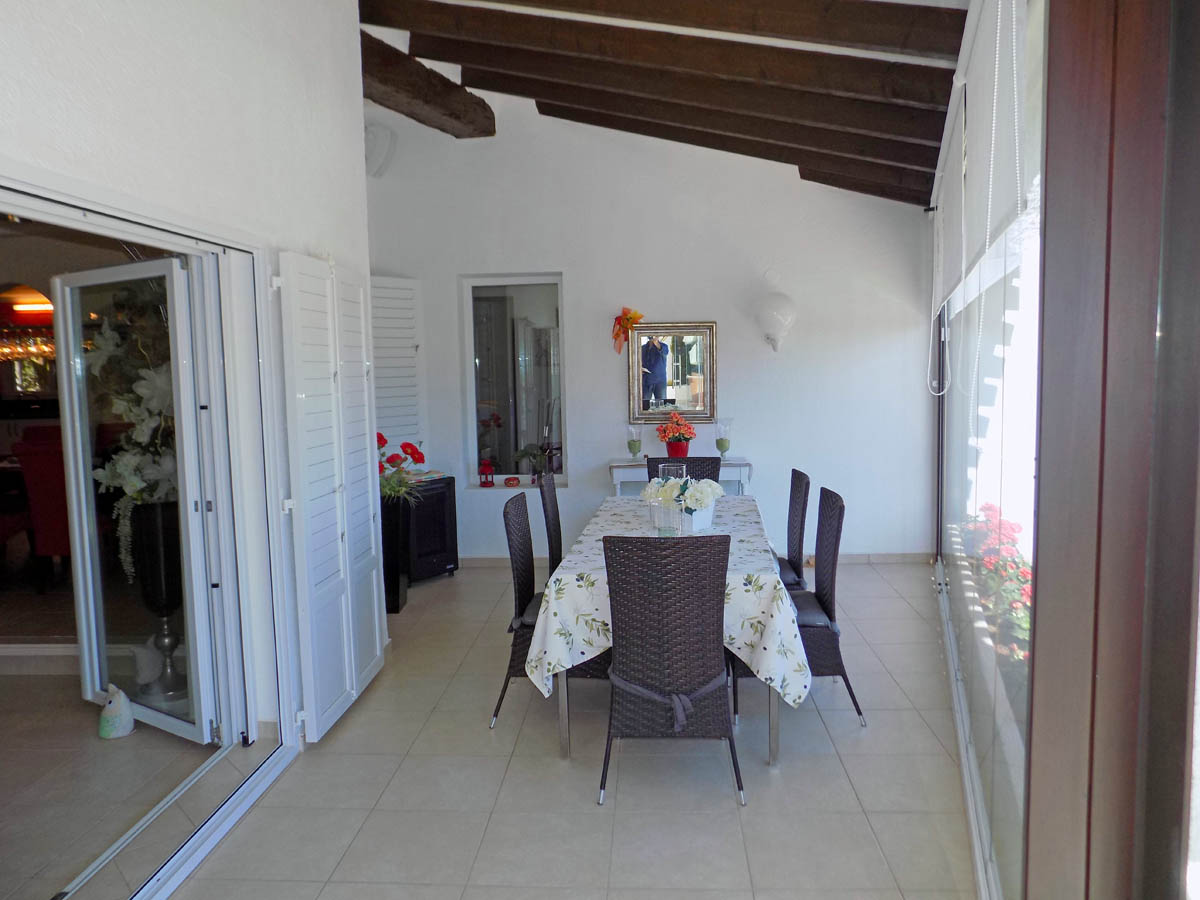 Terraza acristalada con techo de vigas de madera vista, zona de comedor con mobiliario de mimbre y acceso directo al interior mediante puertas blancas.