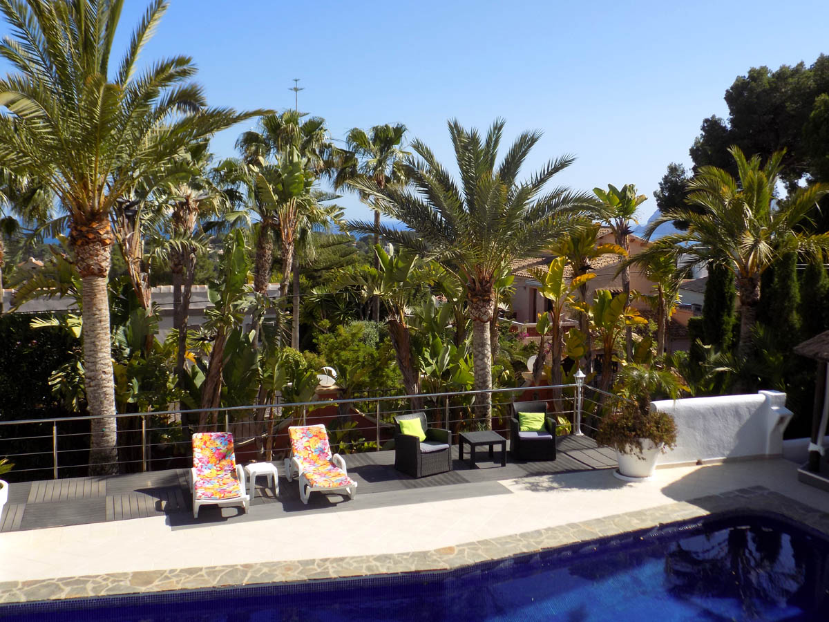 Terraza de madera junto a la piscina de 12x5,5m, rodeada de palmeras maduras y vegetación mediterránea con vistas despejadas hacia la costa de Moraira.
