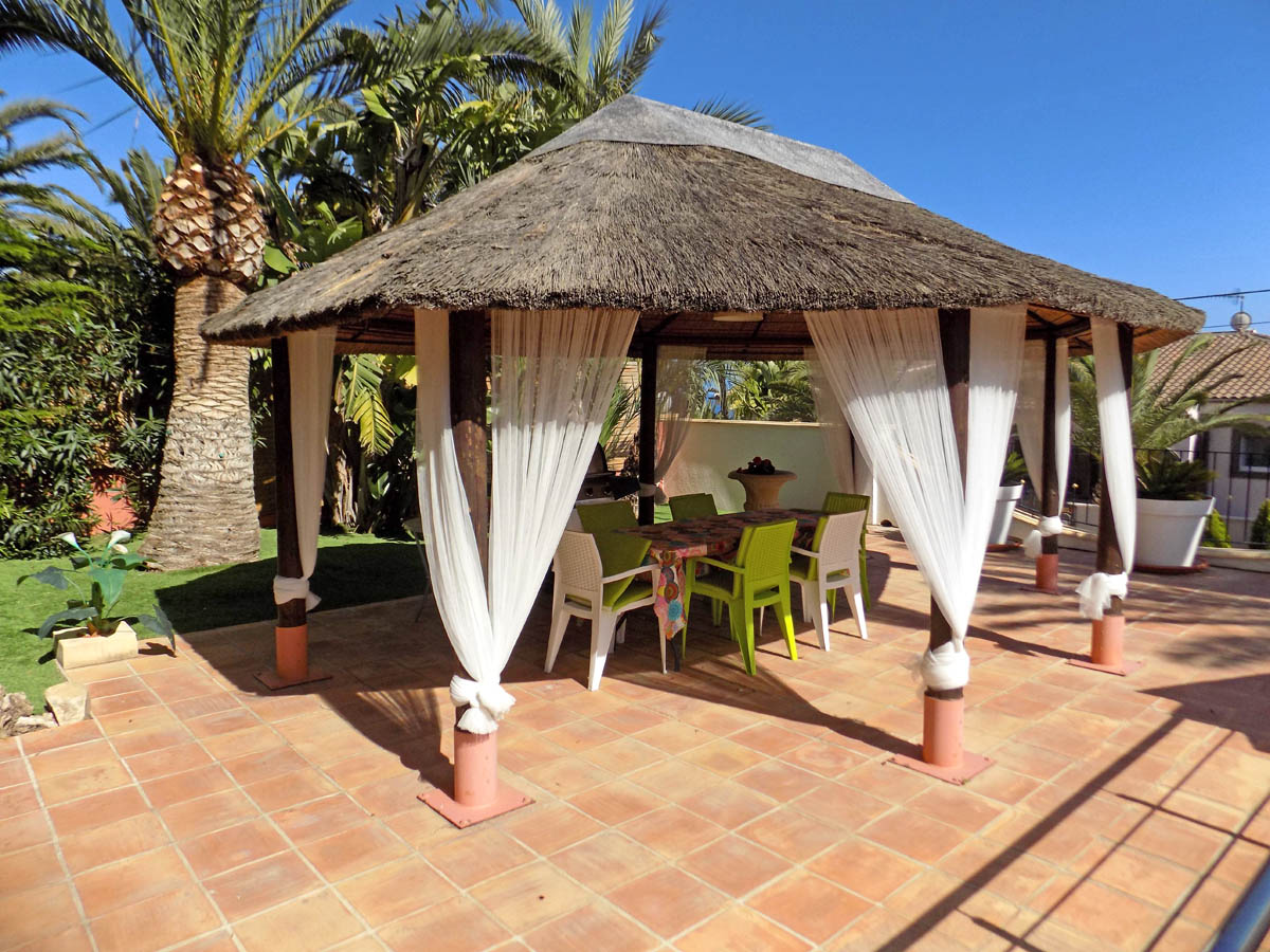 Zona chill-out exterior con cenador de brezo, cortinas blancas decorativas y comedor rodeado de vegetación mediterránea y palmeras.