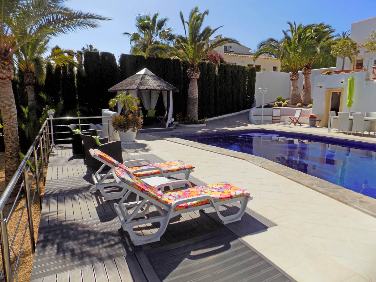 Zona de piscina con solárium de gres y tarima, tumbonas, cenador balinés con techo de paja y jardines maduros con palmeras y setos de privacidad.