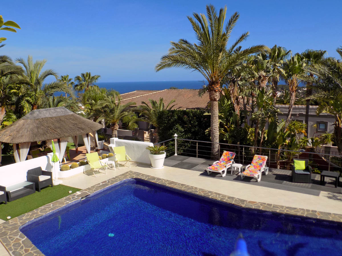 Zona de piscina con terraza solárium, cenador de estilo tropical con techo de paja y vistas panorámicas al mar rodeada de palmeras y vegetación.