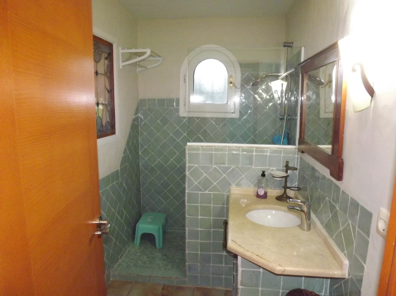 Baño con azulejos verdes en disposición diagonal, encimera de mármol con lavabo integrado y una ducha de obra con ventana de arco.