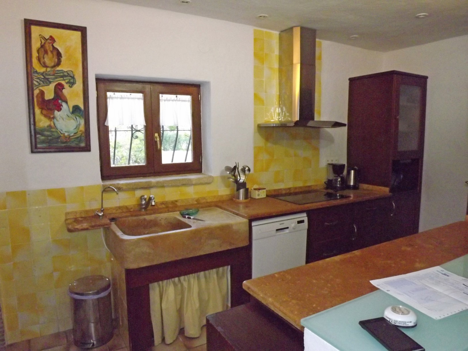 Cocina rústica con fregadero de piedra tallada, azulejos amarillos y armarios de madera oscura. Incluye encimera de mármol, campana de acero y ventana con marco de madera.