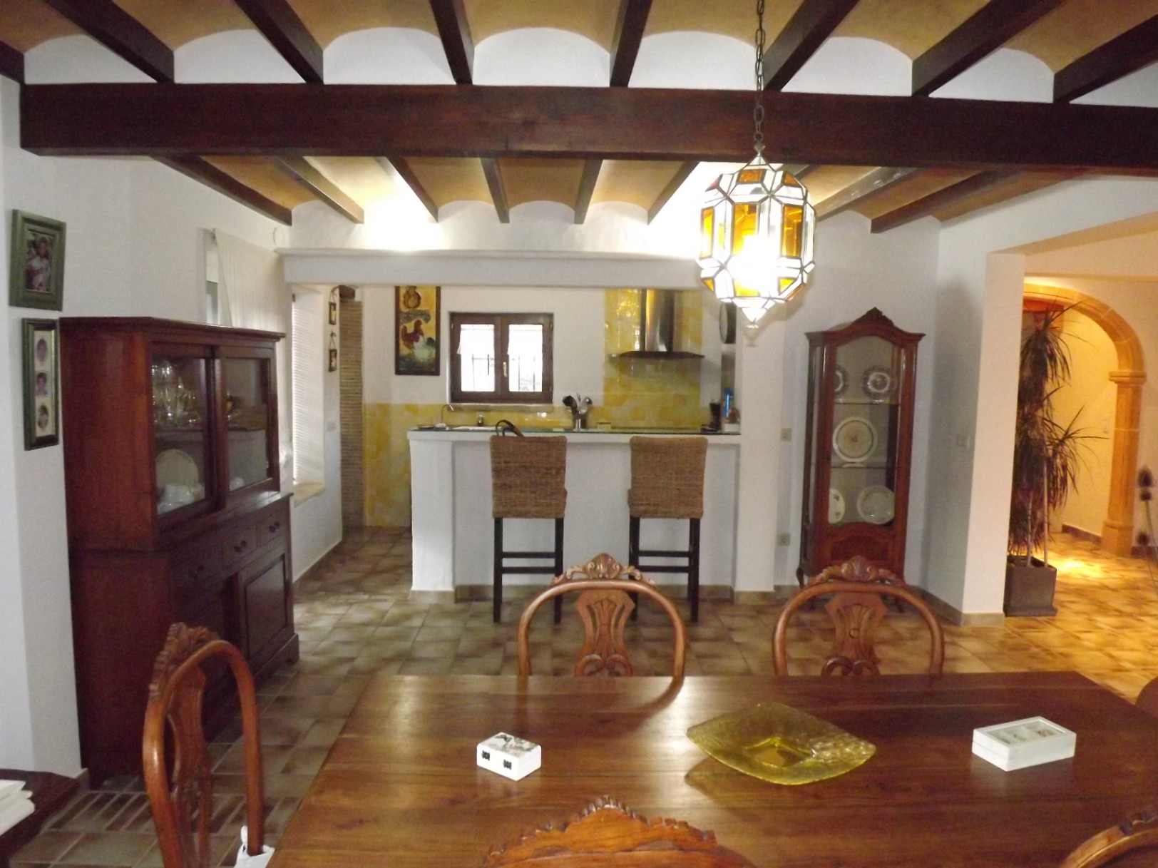 Comedor con vigas de madera expuestas y techo abovedado, conectado a una cocina abierta con barra americana, taburetes de mimbre y suelos cerámicos tradicionales.