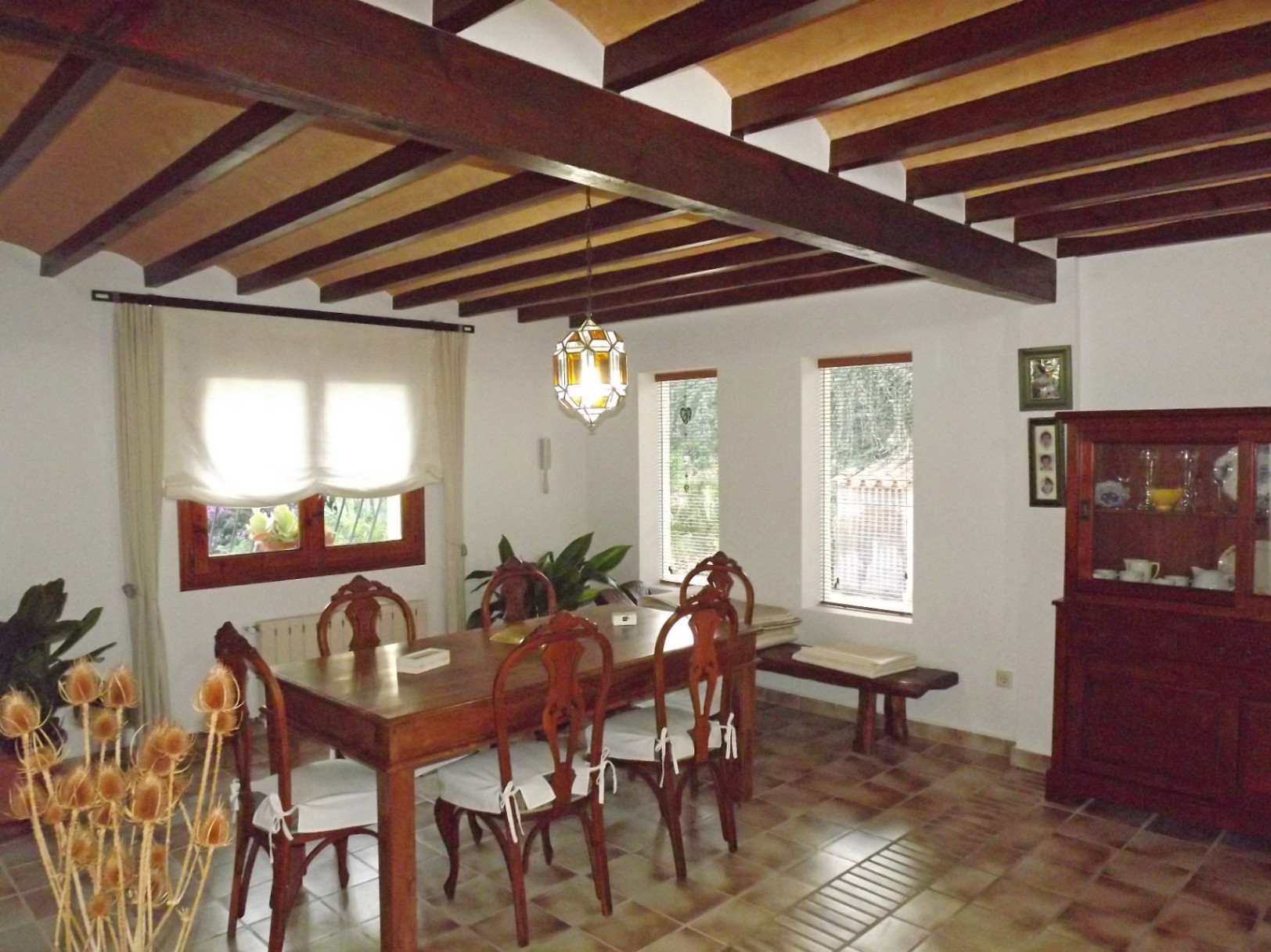 Comedor de estilo rústico con vigas de madera oscura, mesa de comedor clásica y suelos cerámicos. La estancia cuenta con luz natural y una vitrina de madera a juego.