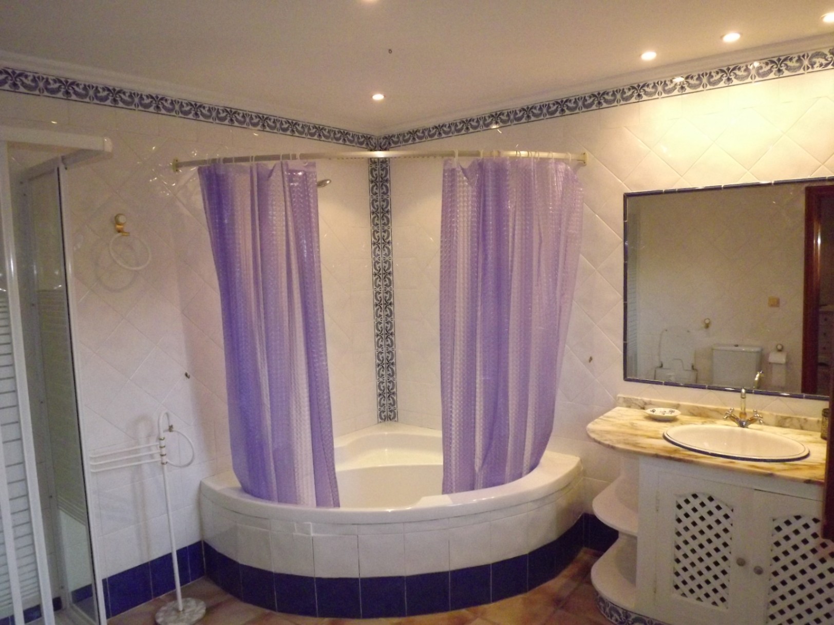 Cuarto de baño con bañera de rincón y cortina púrpura, encimera de mármol con mueble de celosía y azulejos blancos con cenefas decorativas azules.