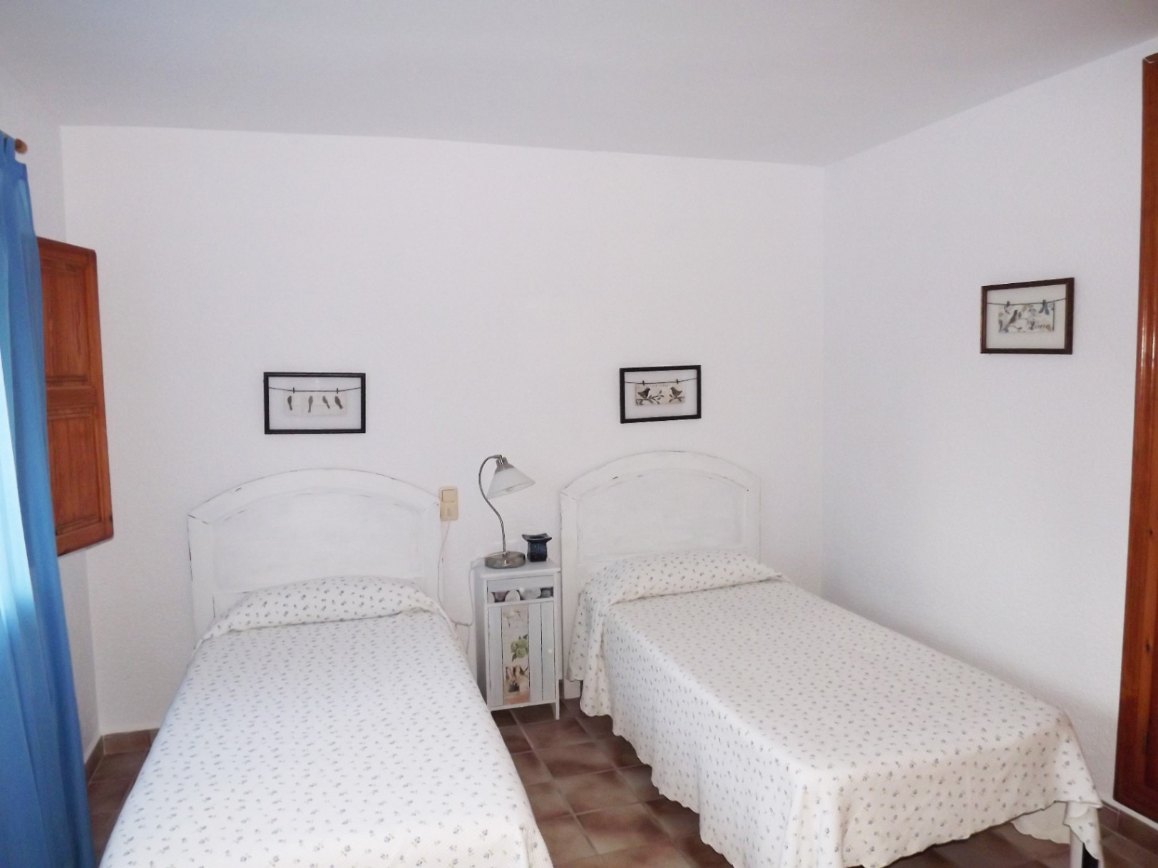 Dormitorio con dos camas individuales, cabeceros blancos de madera y suelo de gres rústico. Incluye contraventanas de madera y luz natural lateral.