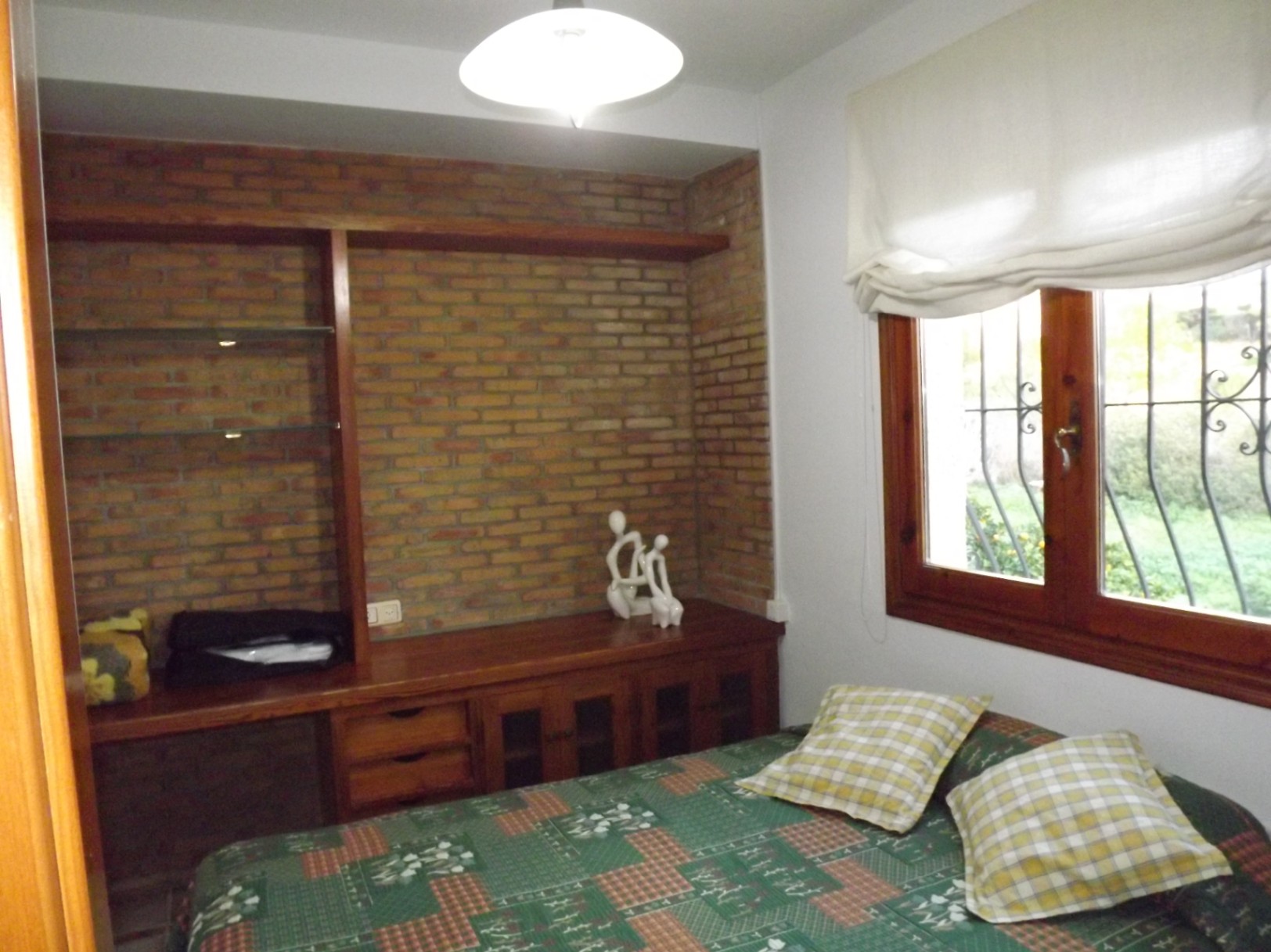 Dormitorio con pared de ladrillo visto, estantería de madera integrada y ventana de madera con vistas al jardín y rejas de seguridad.