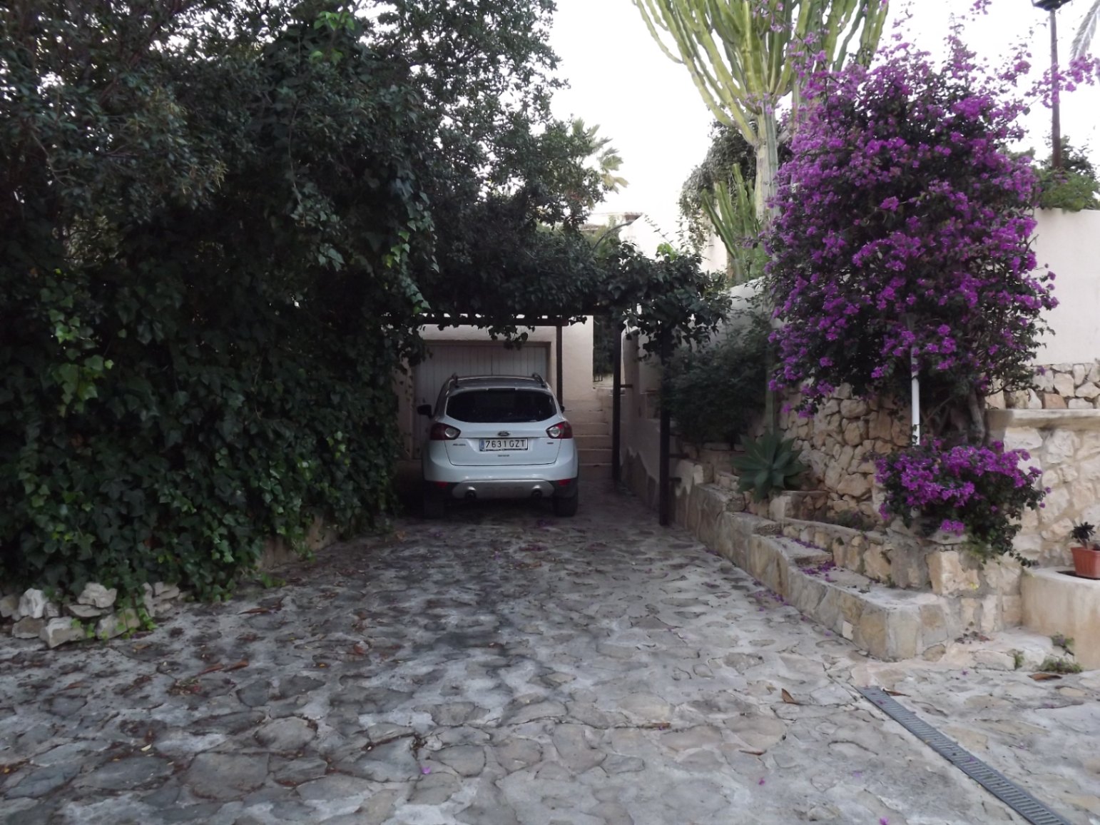 Entrada empedrada con zona de aparcamiento cubierta por vegetación, muros de piedra natural y buganvillas en flor que enmarcan el acceso al garaje.