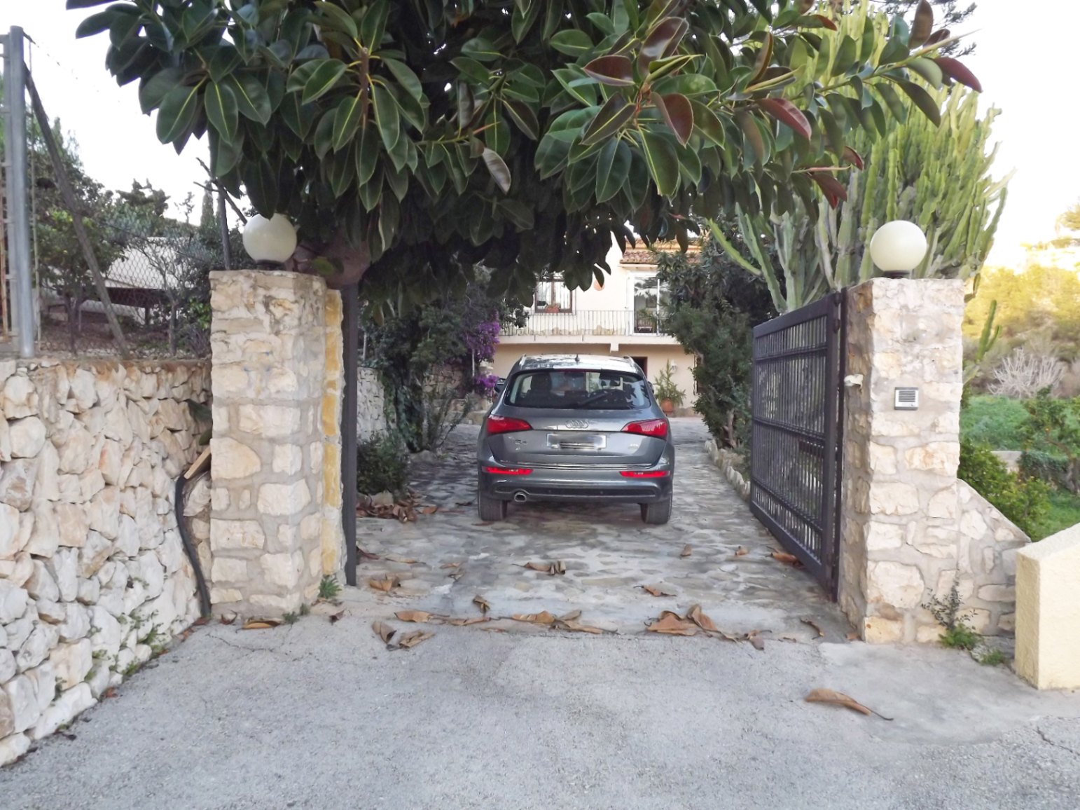 Entrada principal con muros de piedra natural, portón metálico y camino pavimentado rodeado de vegetación mediterránea y árboles maduros.
