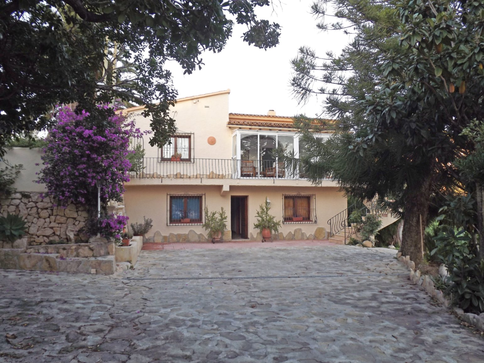 Fachada principal de la villa con patio de piedra natural, terraza acristalada en la planta superior y vegetación mediterránea madura.