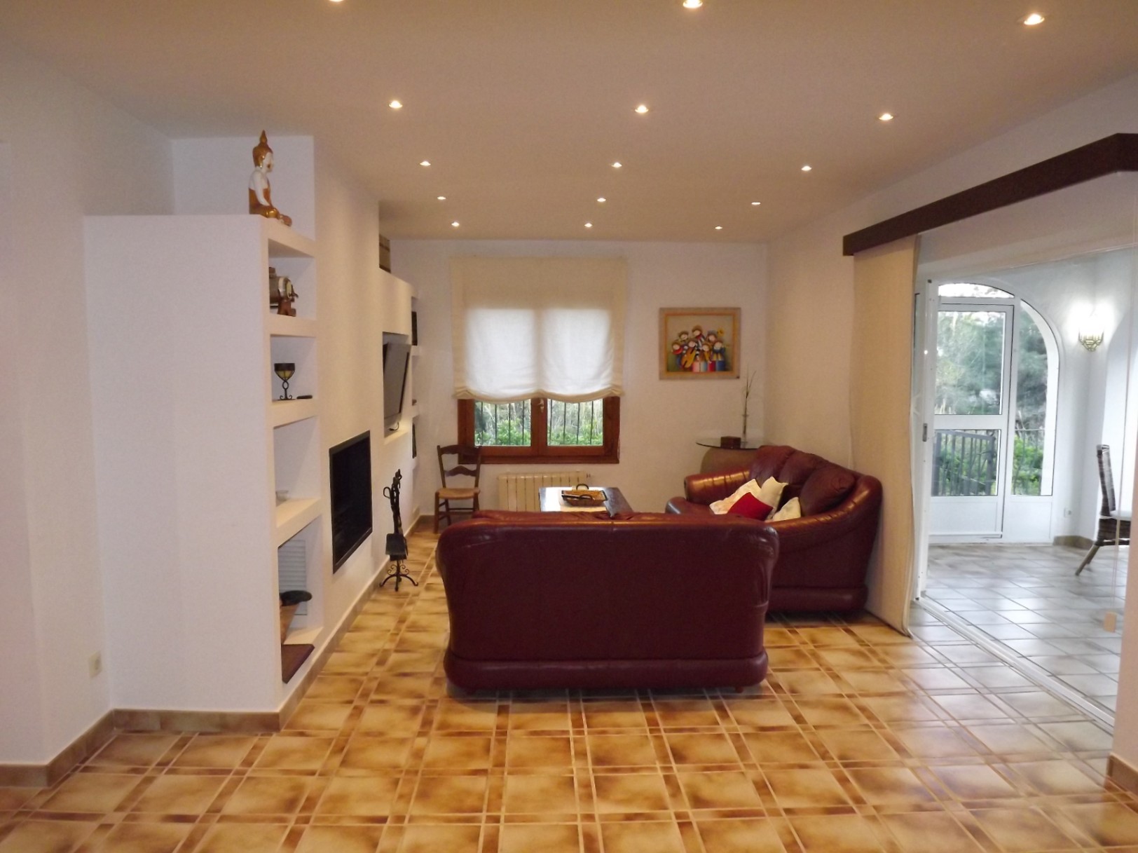 Salón acogedor con chimenea integrada, sofás de cuero burdeos y suelos cerámicos. La estancia cuenta con iluminación empotrada y acceso directo a la terraza acristalada.