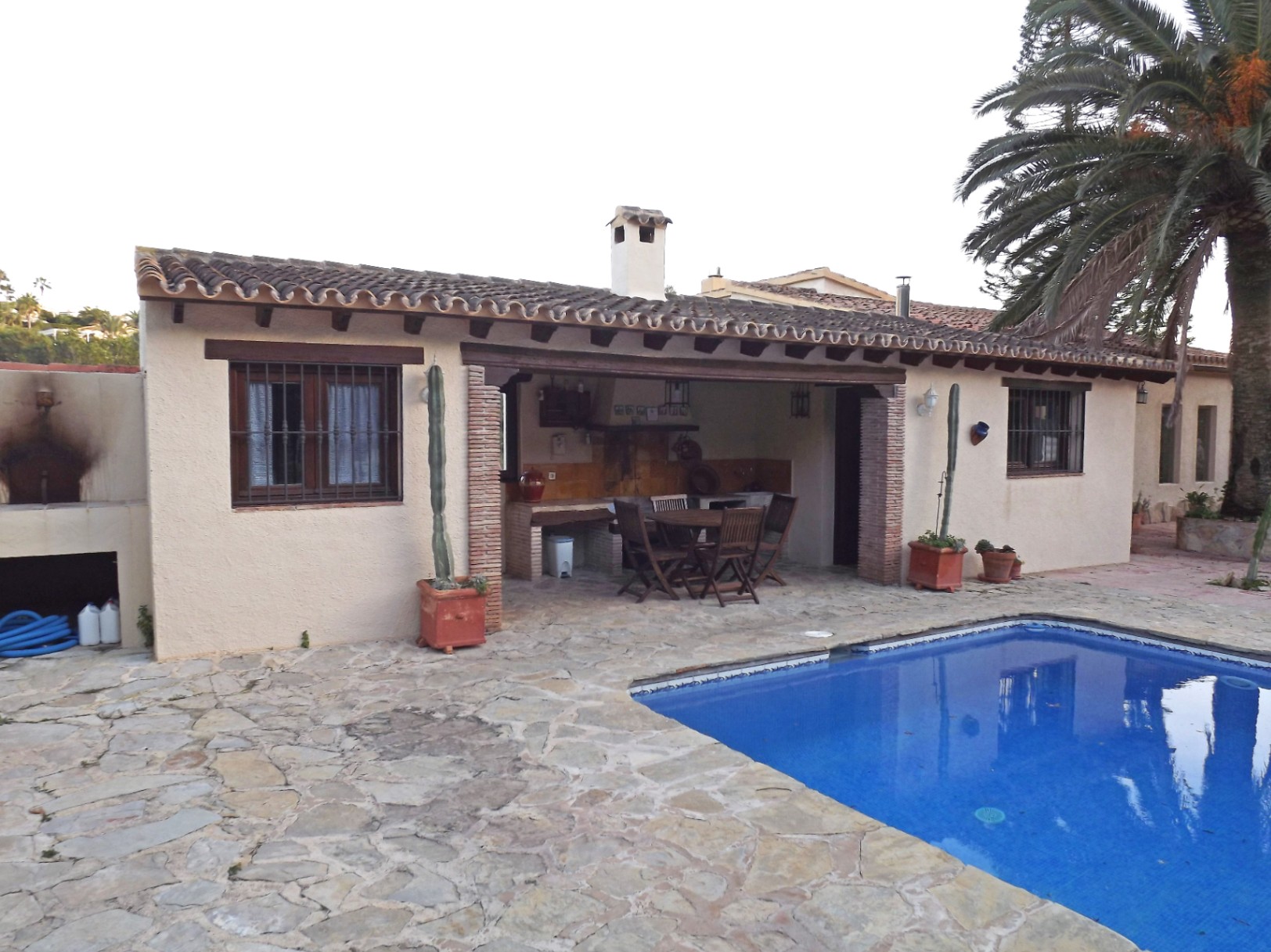 Terraza exterior con suelo de piedra natural, piscina de 12x5m y zona de barbacoa cubierta con vigas de madera y comedor de exterior junto a una palmera.
