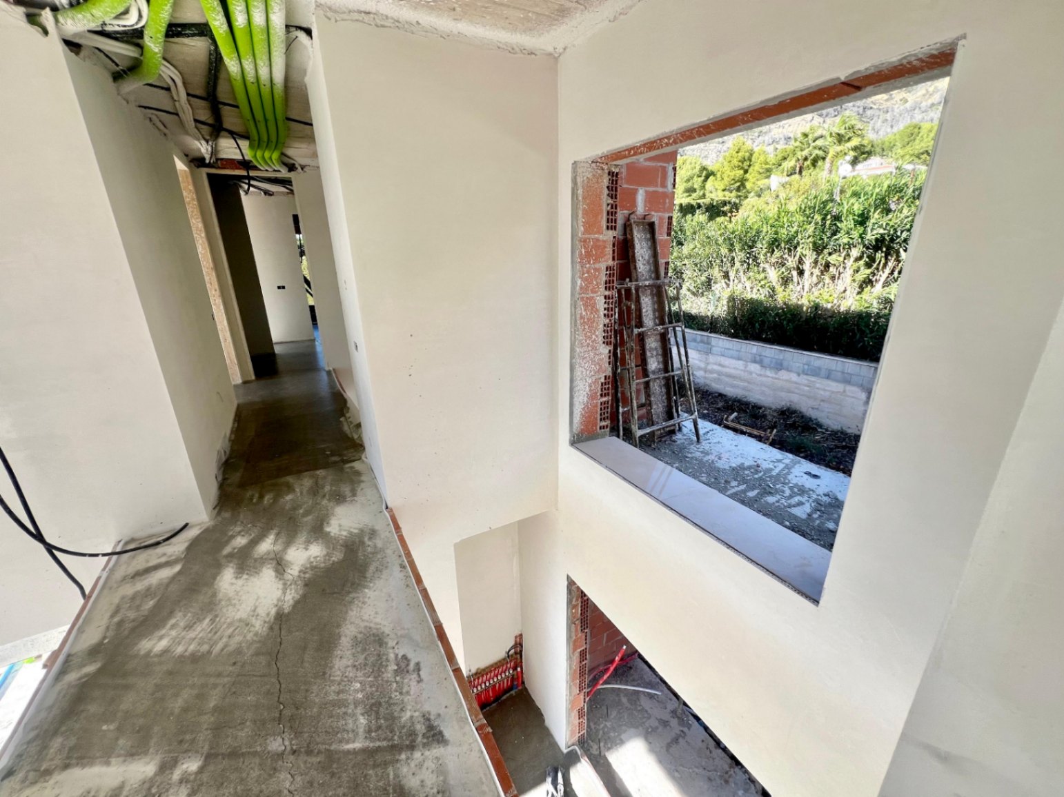 Vista del pasillo interior en fase de construcción, con instalaciones técnicas visibles y un gran hueco para ventanal con vistas al jardín y la montaña.