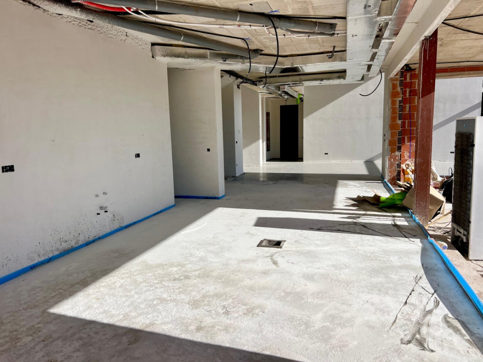 Vista interior de la planta principal en construcción, mostrando techos con instalaciones técnicas, paredes enlucidas y suelos de hormigón con luz natural lateral.