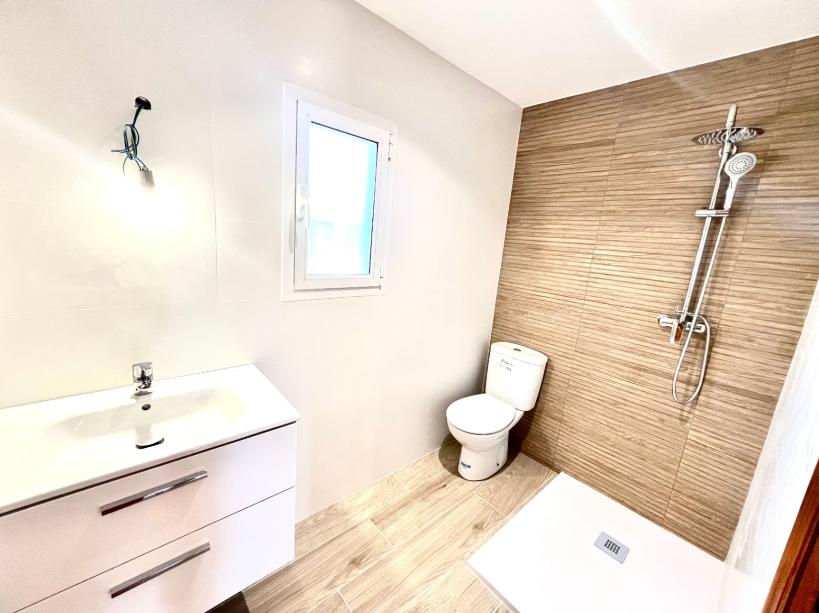 Baño moderno con ducha a ras de suelo, azulejos con textura de madera, mueble de lavabo blanco y ventana para ventilación natural.