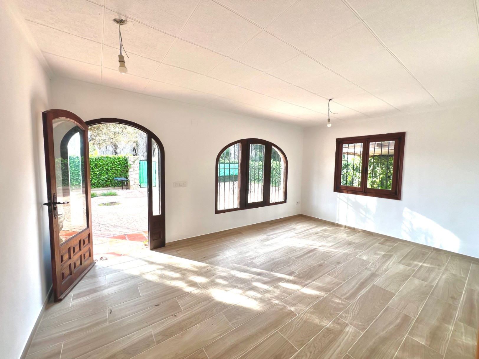 Estancia luminosa con suelos de gres imitación madera, ventanas de madera en arco con rejas de seguridad y acceso directo al patio exterior mediante una puerta acristalada.