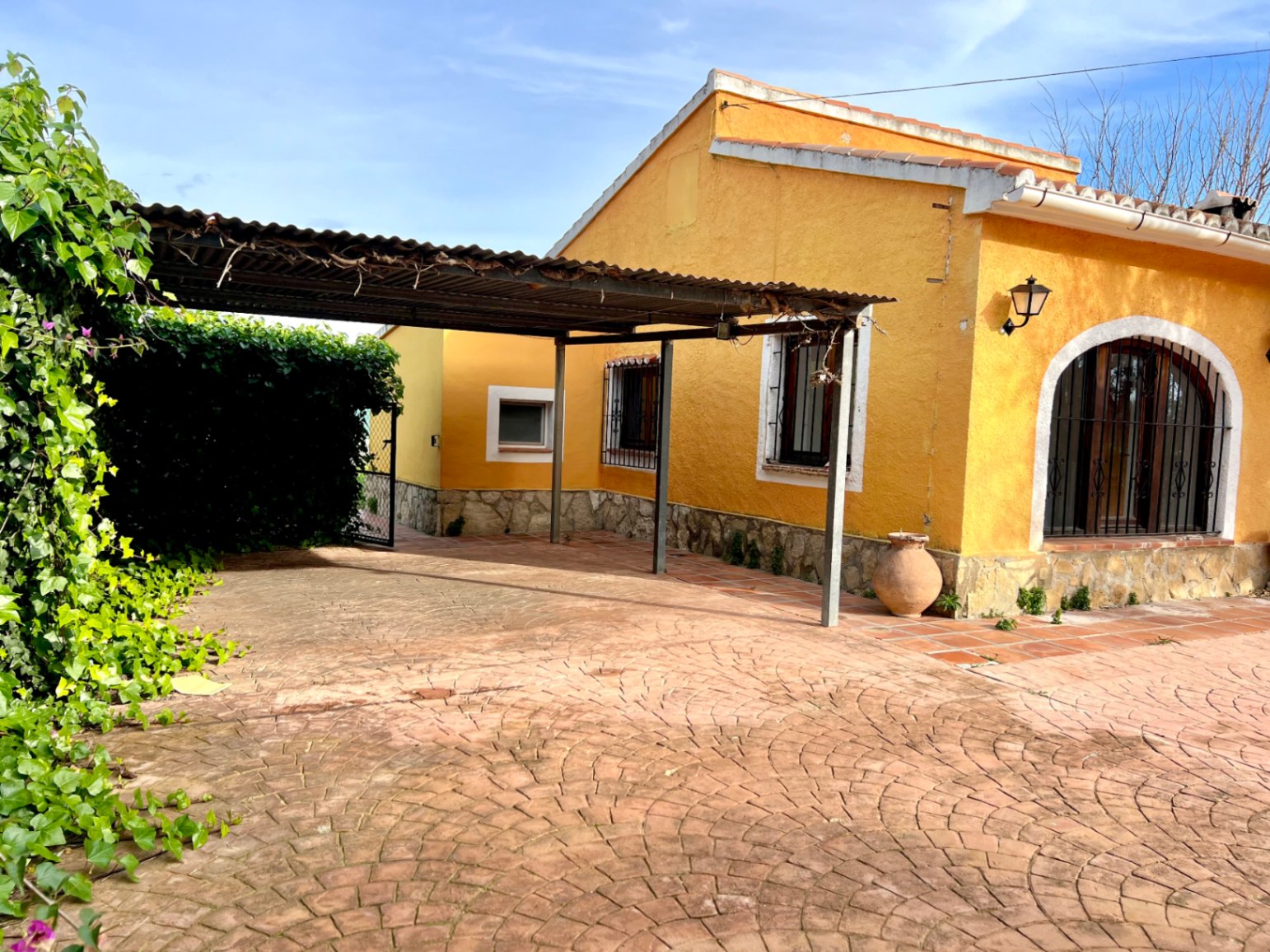 Fachada de color amarillo con zócalo de piedra natural, zona de aparcamiento cubierto y suelo pavimentado con diseño de abanico junto a un ventanal de arco.