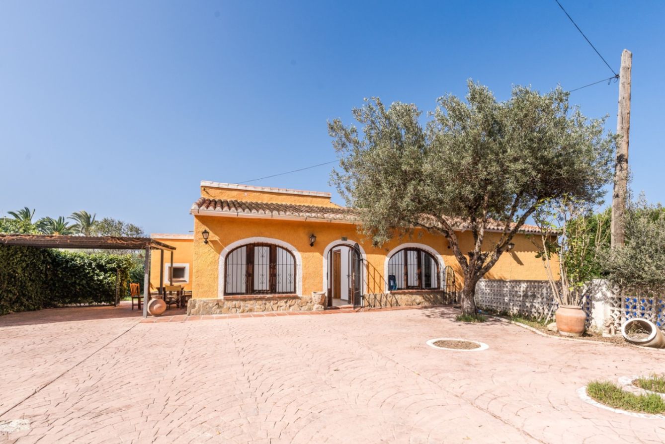 Fachada de estilo mediterráneo con muros ocres, ventanas de arco y zócalo de piedra natural, frente a un amplio patio pavimentado con un olivo centenario.