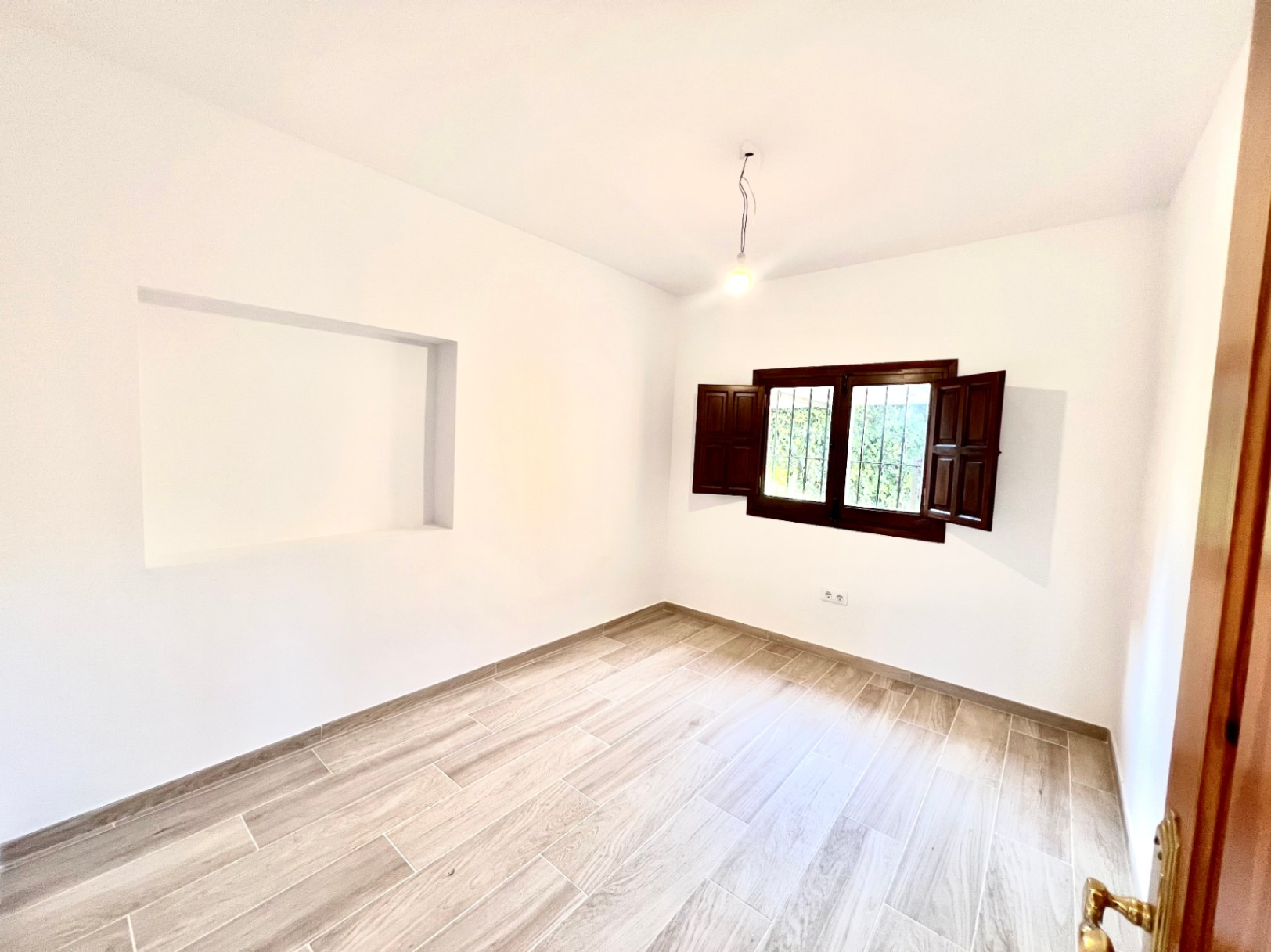 Habitación luminosa con suelos de gres efecto madera, paredes blancas con hornacina decorativa y ventana de madera oscura con contraventanas tradicionales.