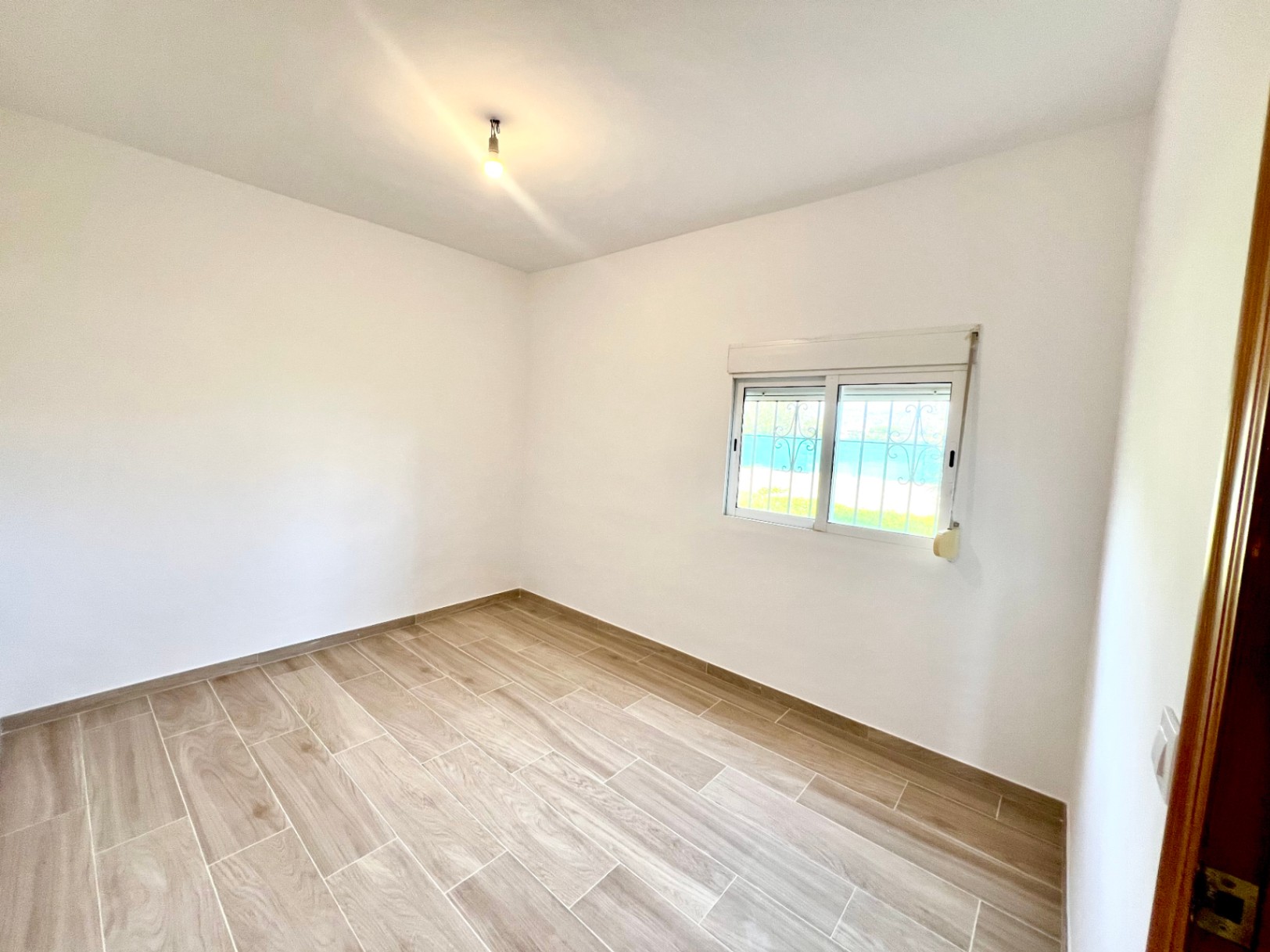 Habitación luminosa con suelos de gres efecto madera, paredes blancas lisas y ventana con reja de seguridad y persiana exterior.