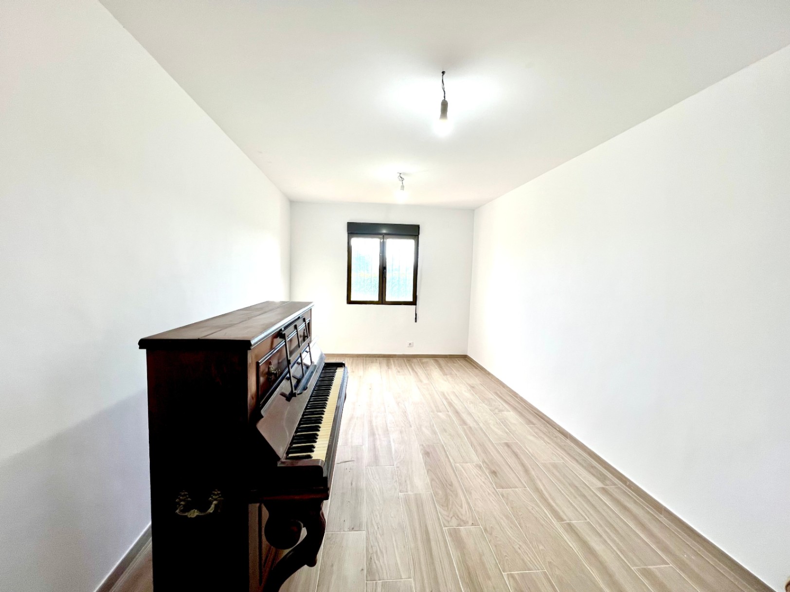 Habitación luminosa de paredes blancas y suelo de gres cerámico imitación madera, equipada con un piano de pared clásico y una ventana de marco oscuro.