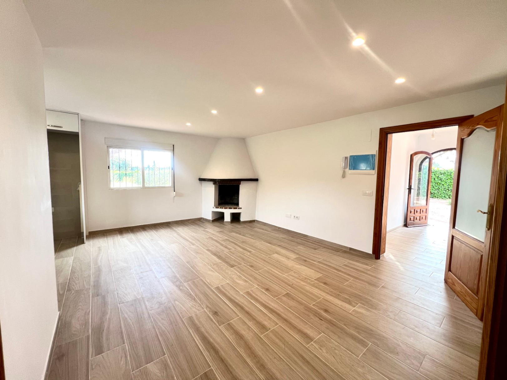 Salón diáfano con chimenea de rincón, suelo de gres cerámico imitación madera y focos empotrados. La estancia conecta con el recibidor a través de carpintería de madera natural.