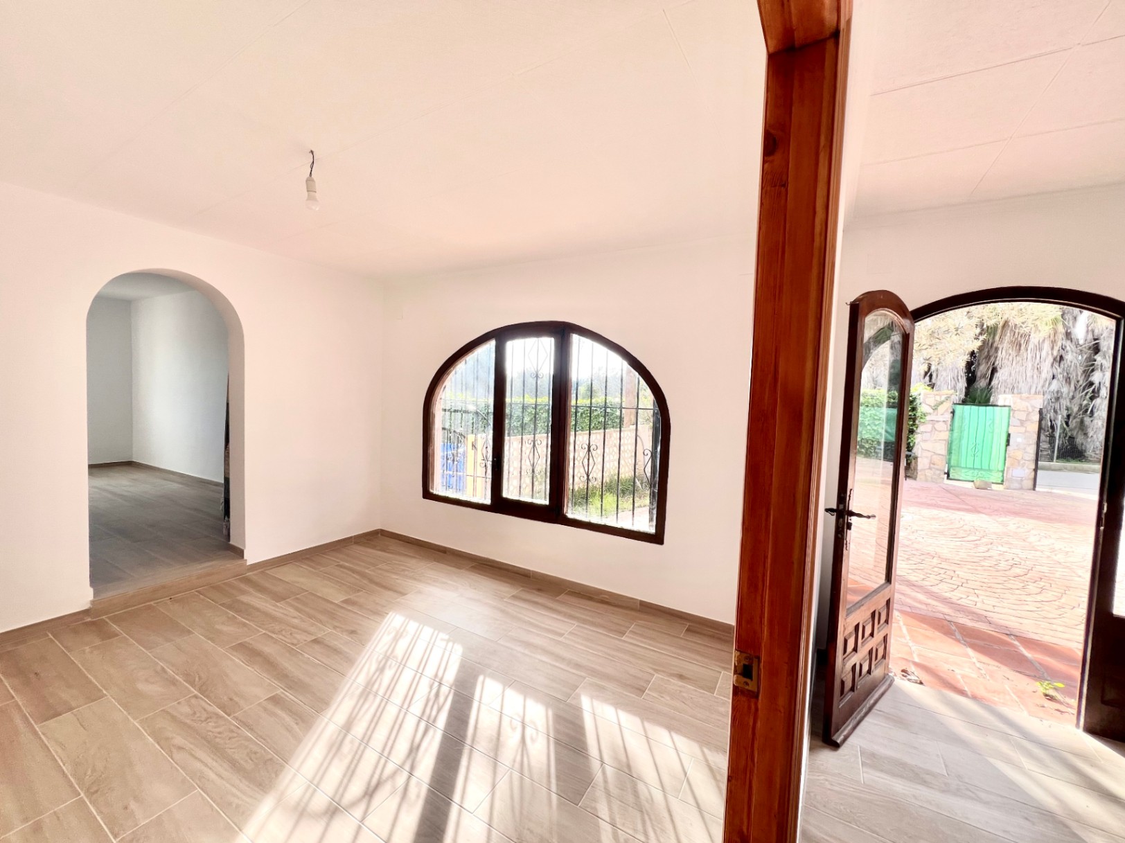 Salón luminoso con suelos de gres efecto madera, ventana de medio punto con marco oscuro y acceso directo al patio exterior pavimentado.