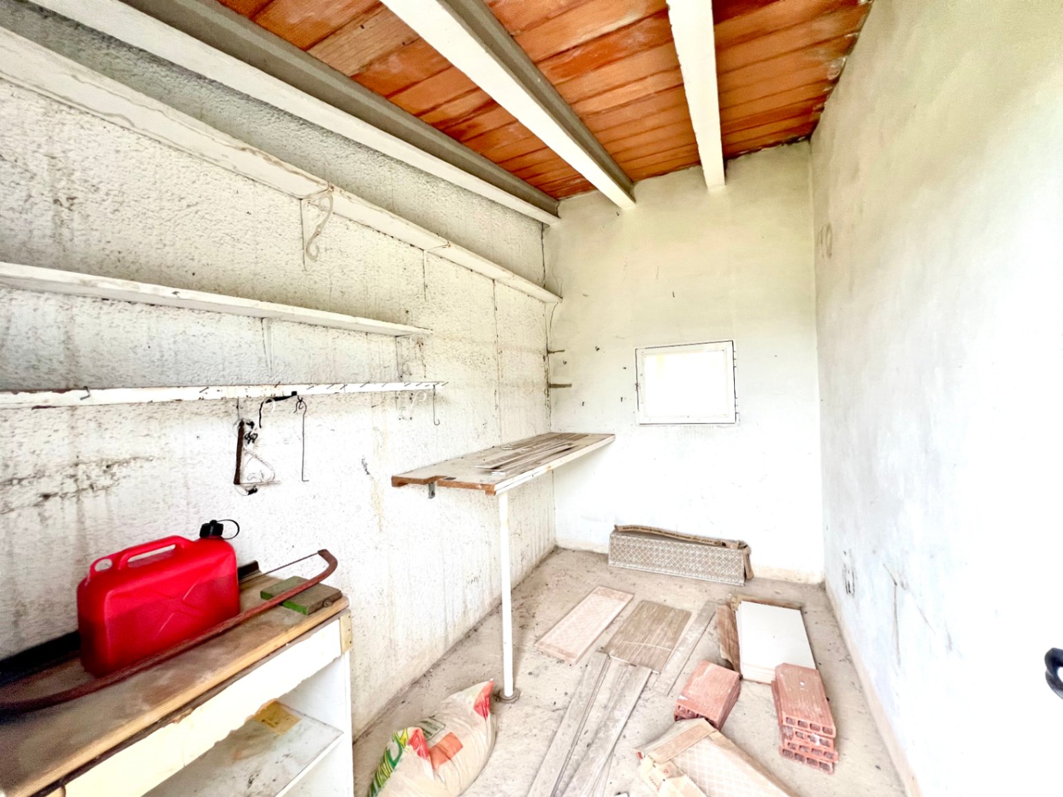 Trastero funcional con techo de vigas de madera y bovedillas, paredes de yeso rugoso, estanterías fijas y luz natural mediante una pequeña ventana.