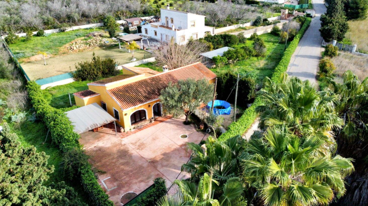 Vista aérea de la vivienda unifamiliar con fachada amarilla y tejado de teja, destacando su amplio patio pavimentado, garaje cubierto y extensa parcela verde.