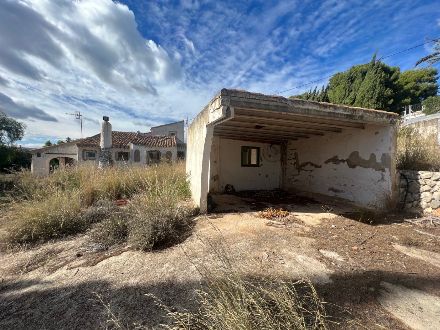Cochera abierta y trastero exterior en estado original, situados en una amplia parcela con vegetación natural y vistas a la vivienda principal de estilo tradicional.