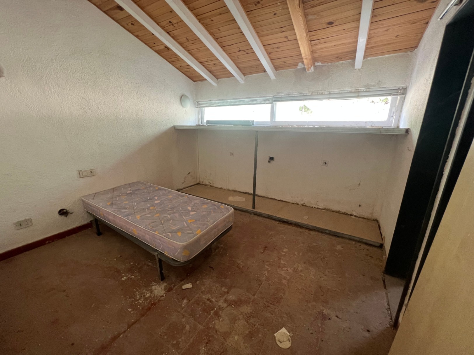 Dormitorio con techo inclinado de vigas de madera expuestas y bovedillas blancas. Incluye suelos de barro originales y una ventana alargada que aporta luz natural.