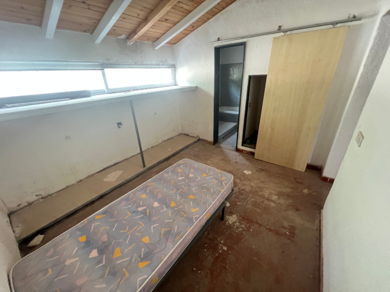 Dormitorio con techo inclinado de vigas de madera, ventana horizontal y puerta corredera que da paso a un baño con azulejos oscuros. Espacio ideal para reforma integral.