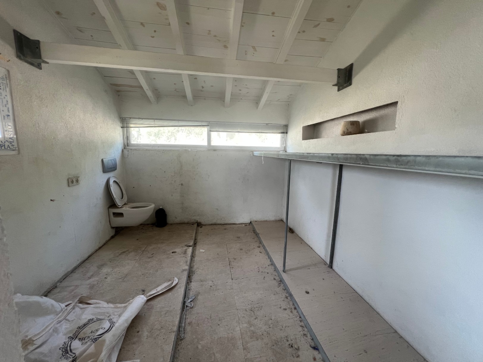 Espacio interior para reformar con techos de vigas de madera blancas, inodoro suspendido y estantería fija. Un ventanal horizontal aporta luz natural a la estancia.