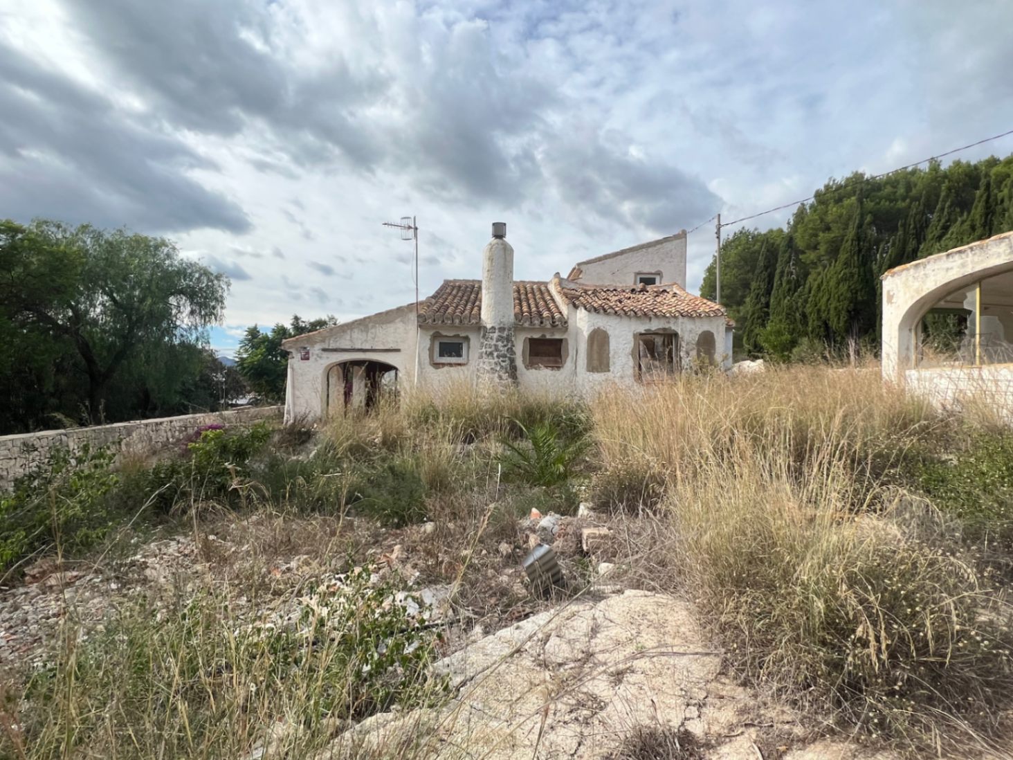 Fachada exterior de chalet rústico en Jávea con chimenea de piedra y arcos tradicionales, rodeado de vegetación natural y parcela extensa para reformar íntegramente.