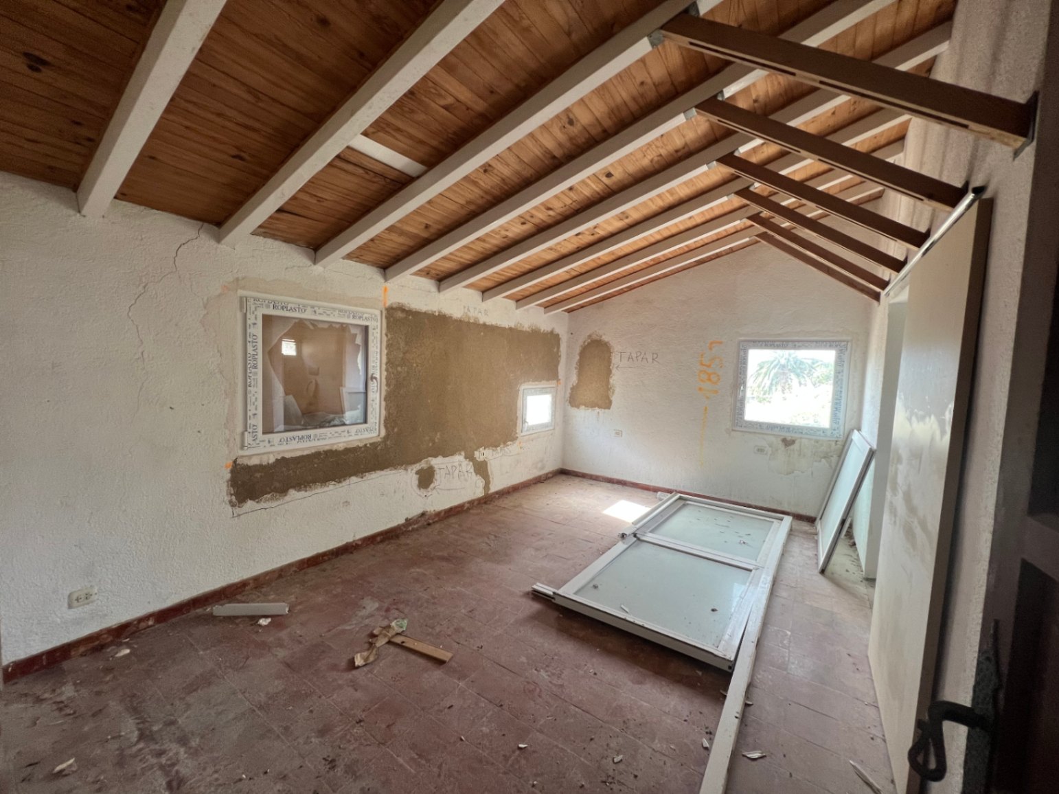 Habitación en fase de reforma con techo inclinado de vigas blancas, suelos de terracota original y nuevas carpinterías de PVC en proceso de instalación.