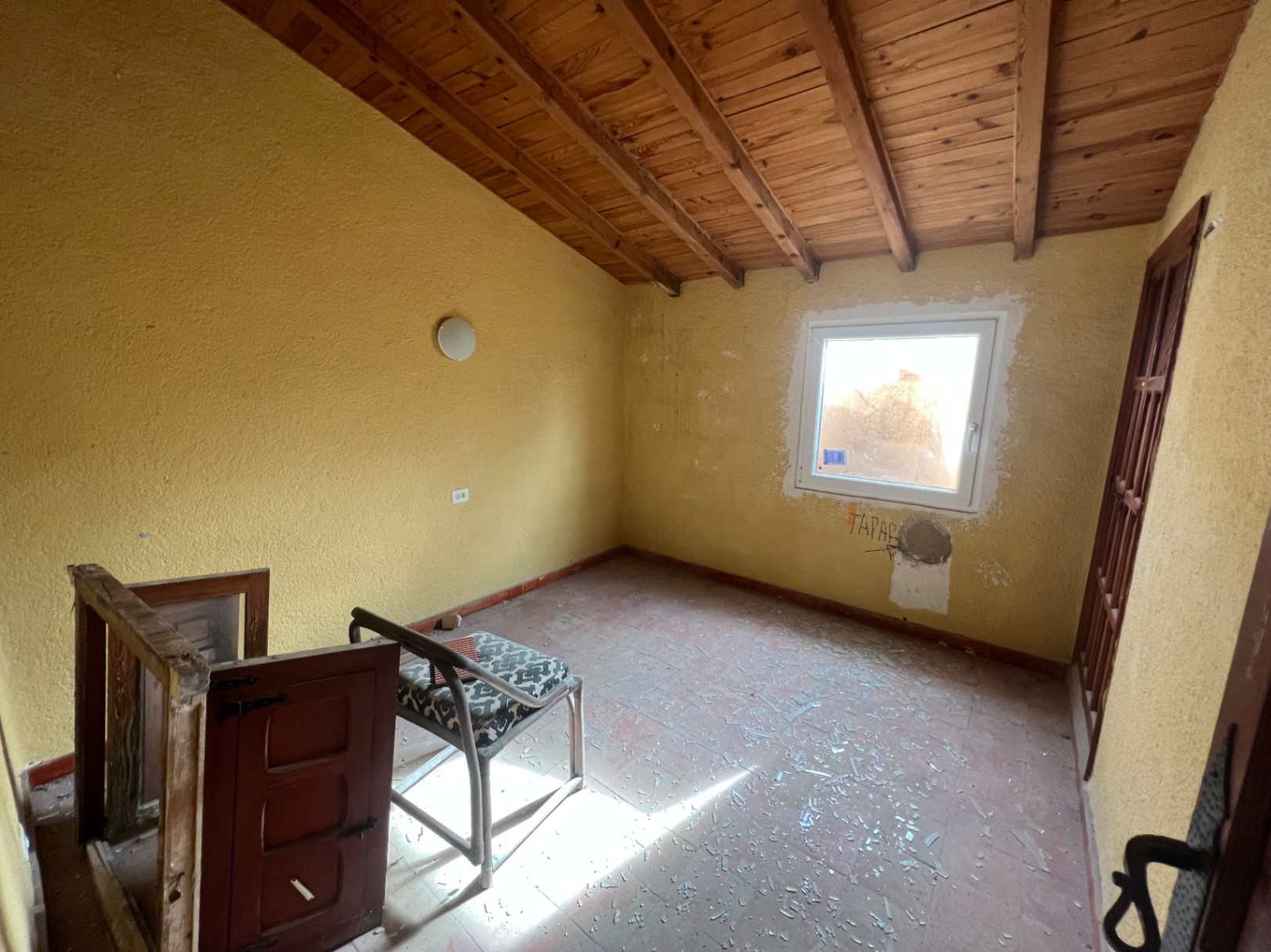 Habitación para reformar con techos inclinados de vigas de madera vistas, suelos de gres y ventana de PVC. Espacio diáfano con gran potencial de personalización.
