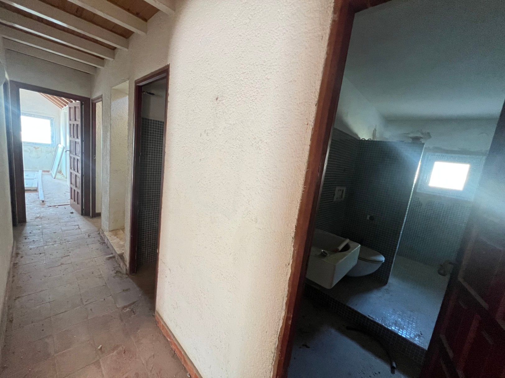 Pasillo interior con vigas de madera vista, suelos cerámicos y acceso a estancias a reformar, incluyendo un baño con azulejos de gresite azul.