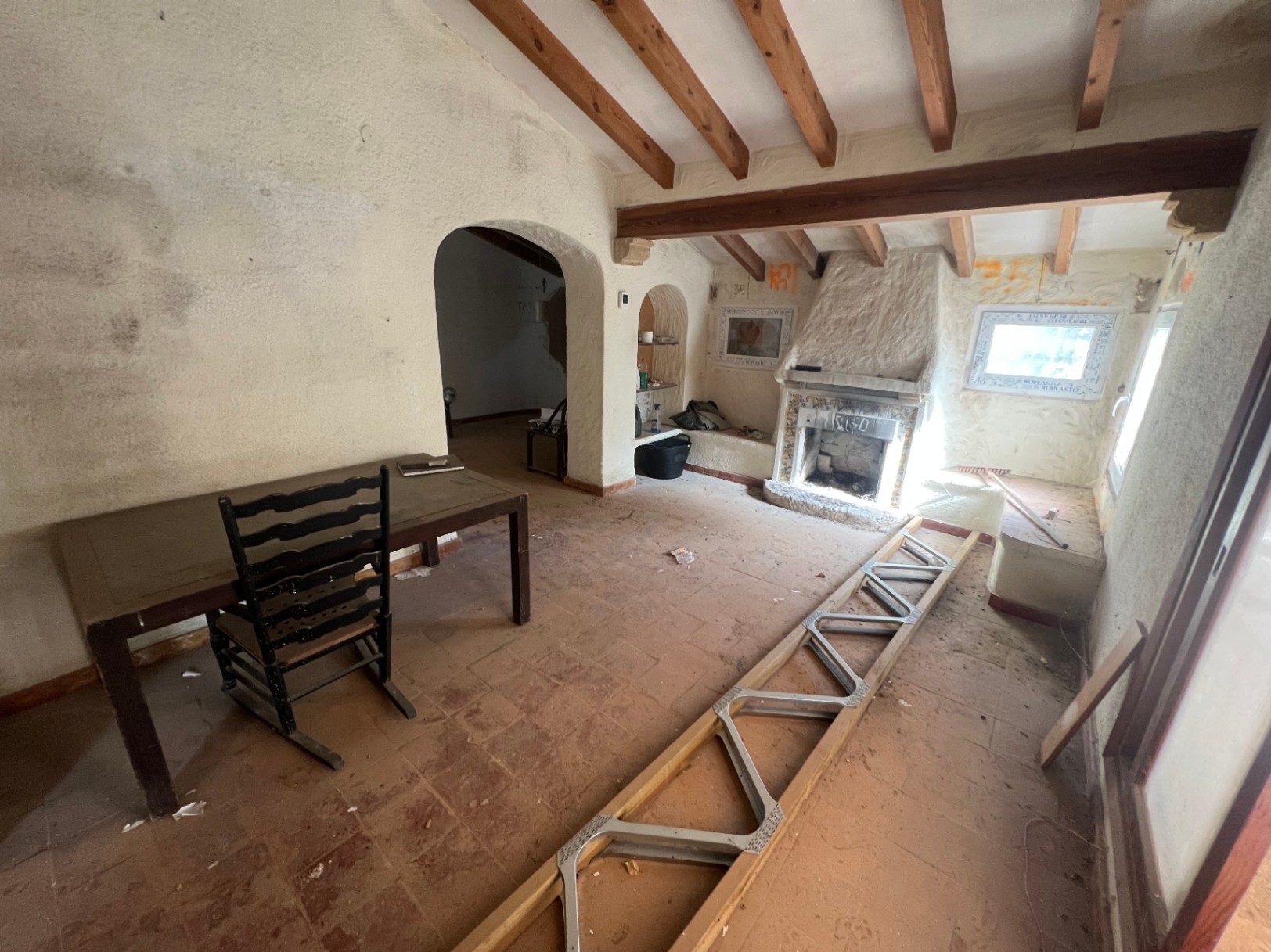 Salón rústico para reformar con vigas de madera a la vista, chimenea tradicional y suelos de barro cocido. Los arcos conectan con otras estancias bajo techos altos.