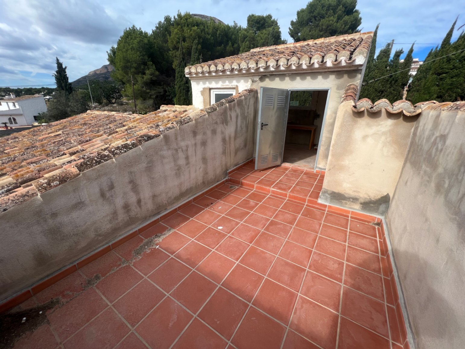 Terraza superior con suelo de baldosas de barro cocido y acceso a un pequeño estudio independiente bajo tejado de teja árabe con vistas al entorno natural.