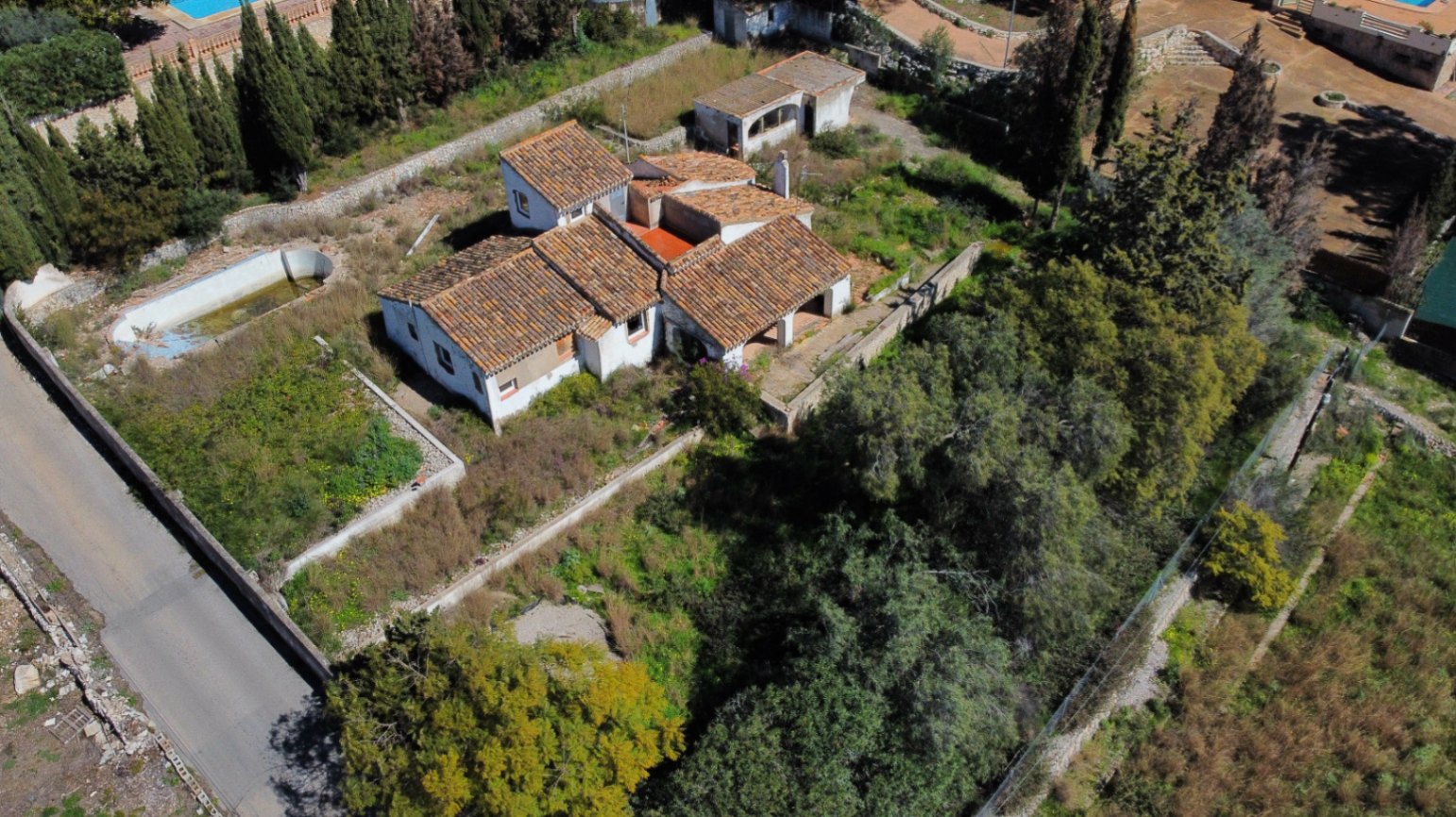 Vista aérea de la propiedad que muestra la estructura de techos de teja, la parcela de 2.895m2 con vegetación autóctona y la piscina de forma arriñonada para reformar.