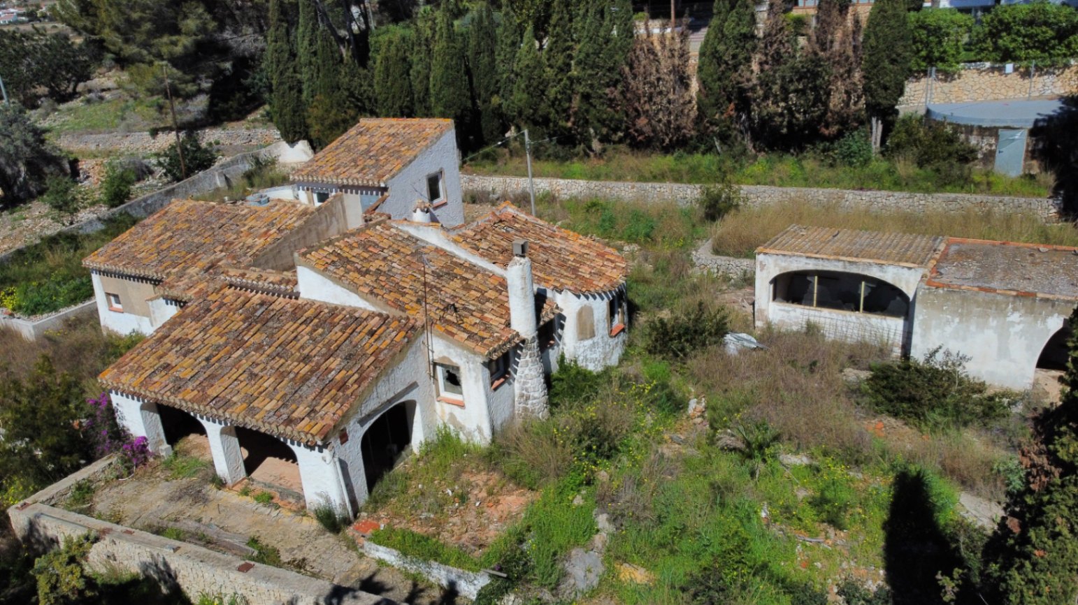 Vista aérea del chalet con tejados de teja árabe, naya de arcos y chimenea de piedra, rodeado de vegetación natural y muros de piedra seca en el Montgó.