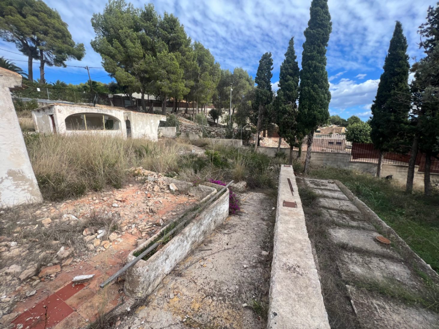 Vista exterior de la parcela con estructuras de hormigón para reformar, rodeada de pinos y cipreses bajo un cielo despejado en la zona del Montgó.
