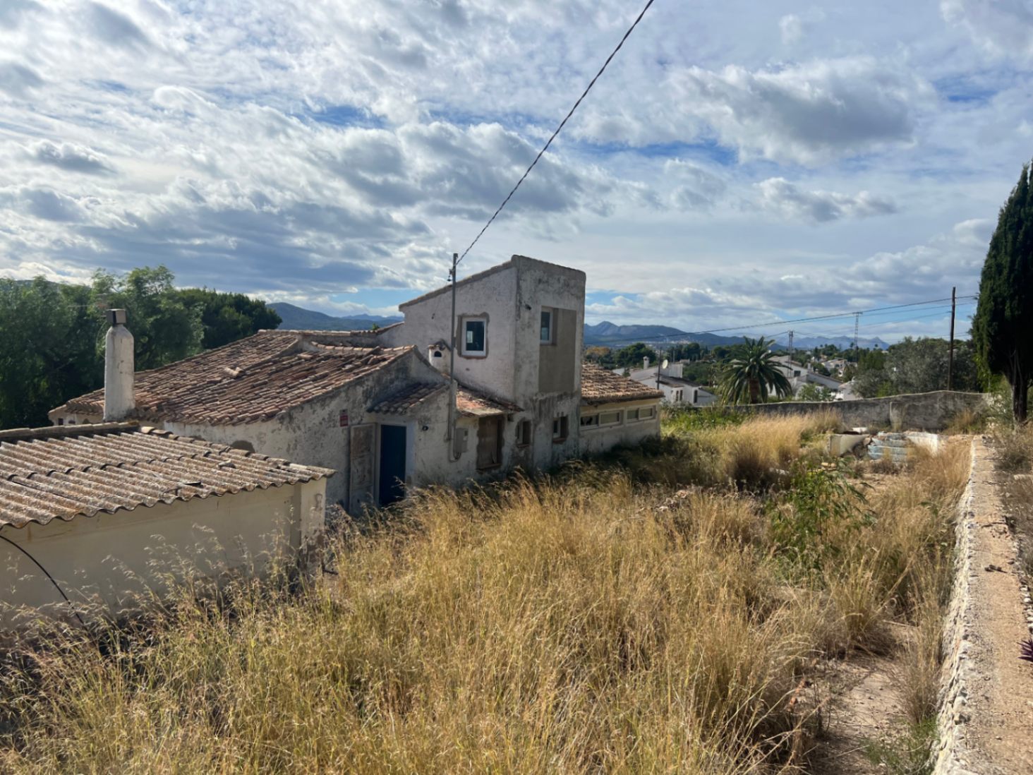 Vista exterior de la vivienda con tejados de teja cerámica y fachada para reformar, situada en una amplia parcela con vegetación natural y vistas a las montañas.