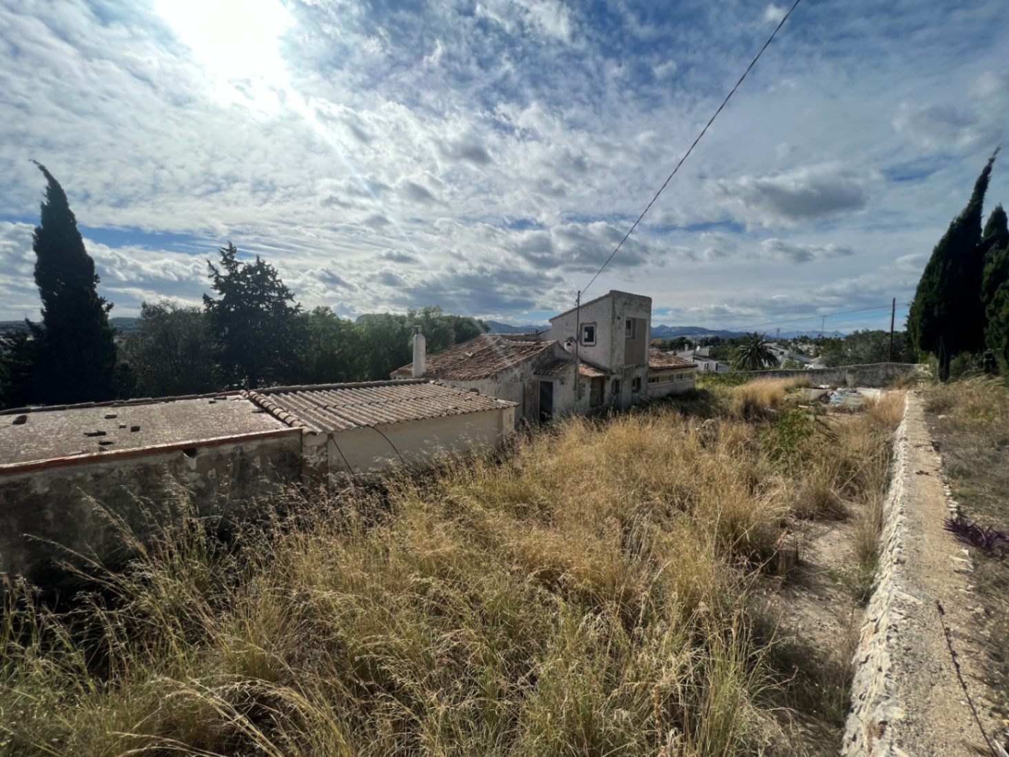 Vista exterior de la vivienda tradicional con tejados de teja árabe y fachada blanca, rodeada de una amplia parcela con vegetación natural y muros de piedra.
