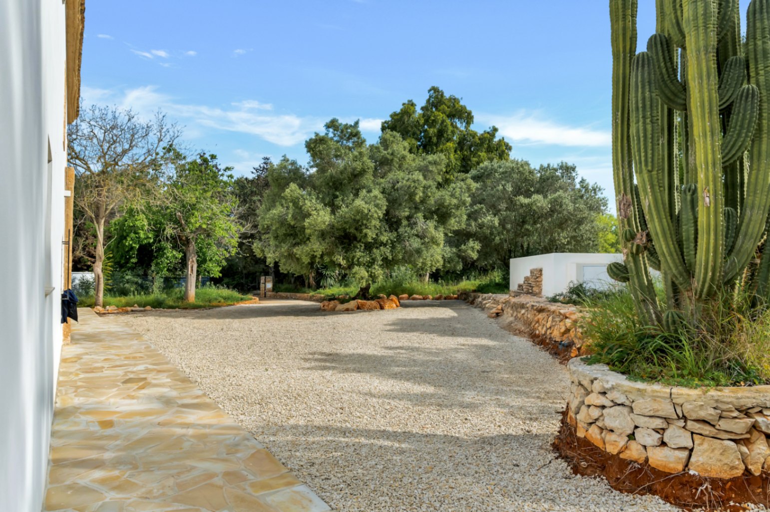 Amplia zona exterior con camino de grava, muros de piedra natural y un gran cactus ornamental junto a la fachada blanca de la vivienda.