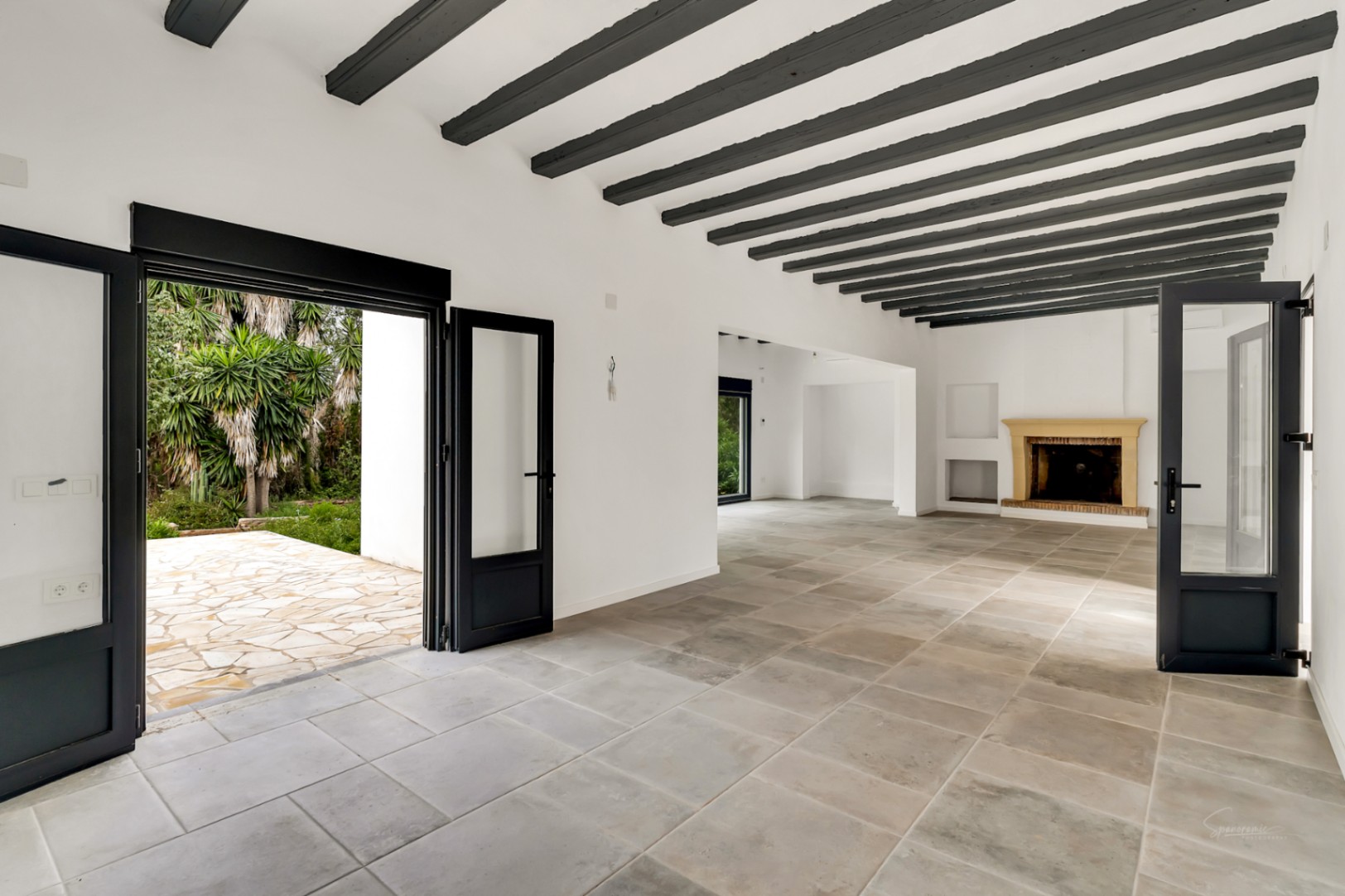 Amplio salón con vigas de madera oscura a la vista, suelos de piedra natural y una chimenea rústica. Las puertas acristaladas conectan el interior con la terraza exterior.
