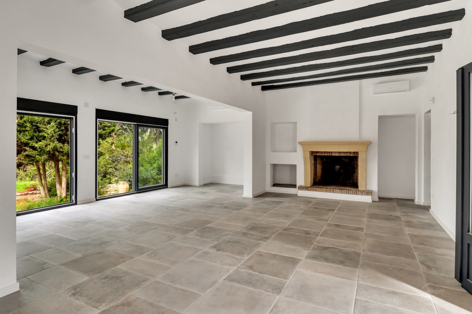 Amplio salón con vigas de madera oscura, chimenea de piedra tradicional y suelos de piedra. Grandes ventanales conectan el interior con el jardín.