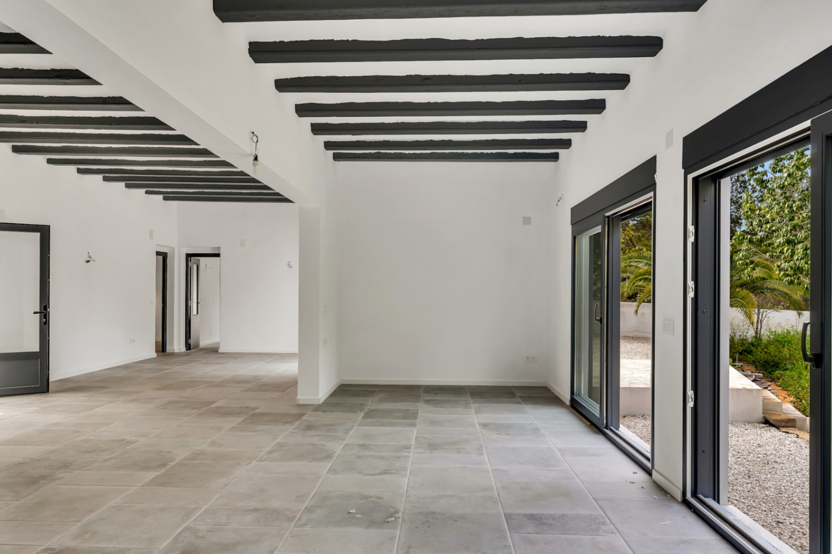 Amplio salón de planta abierta con vigas de madera oscura expuestas, suelos de gres porcelánico gris y grandes ventanales correderos que conectan con el jardín exterior.