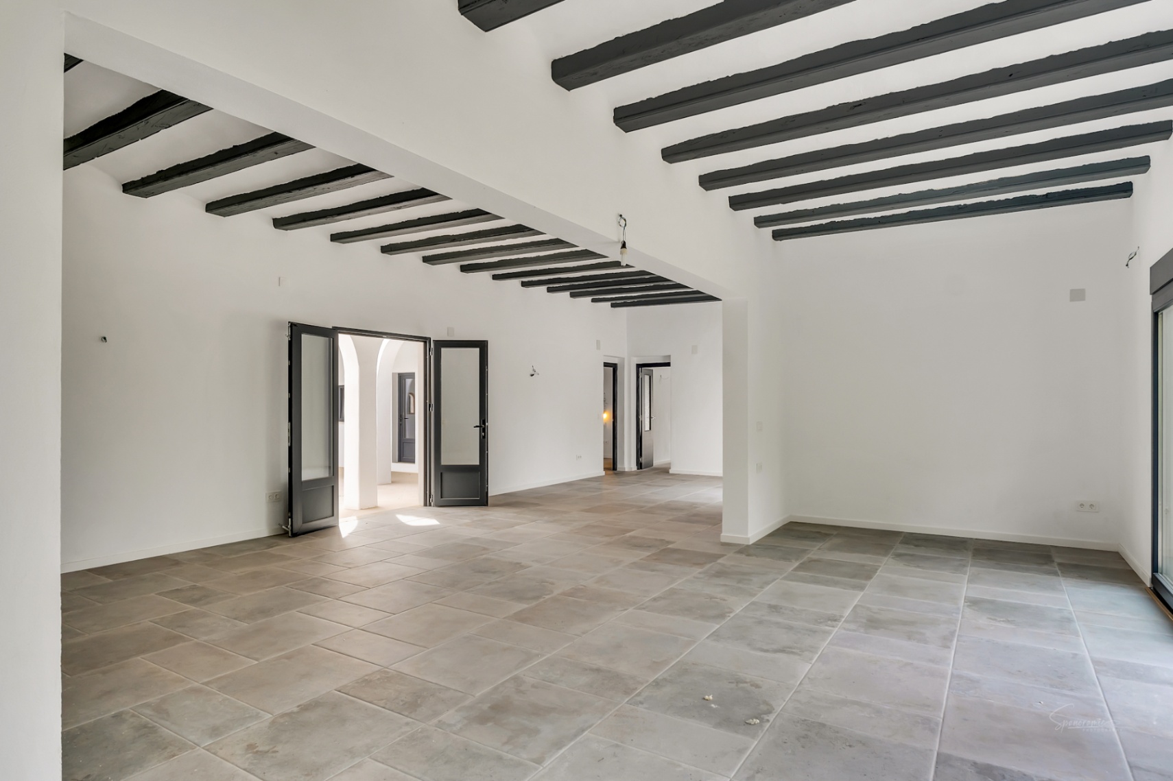 Amplio salón diáfano con vigas de madera oscura expuestas, suelos de piedra de gran formato y acceso directo a un patio interior con arcos tradicionales.